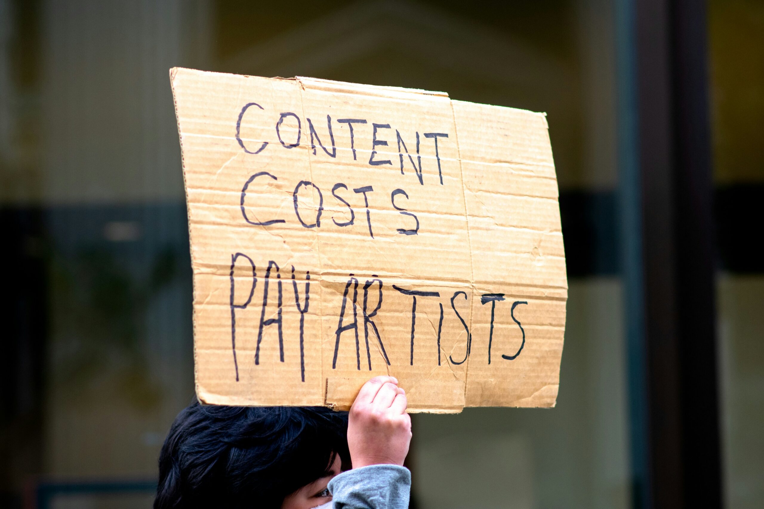 Person holding a sign that says 'Content Costs, Pay Artists'.