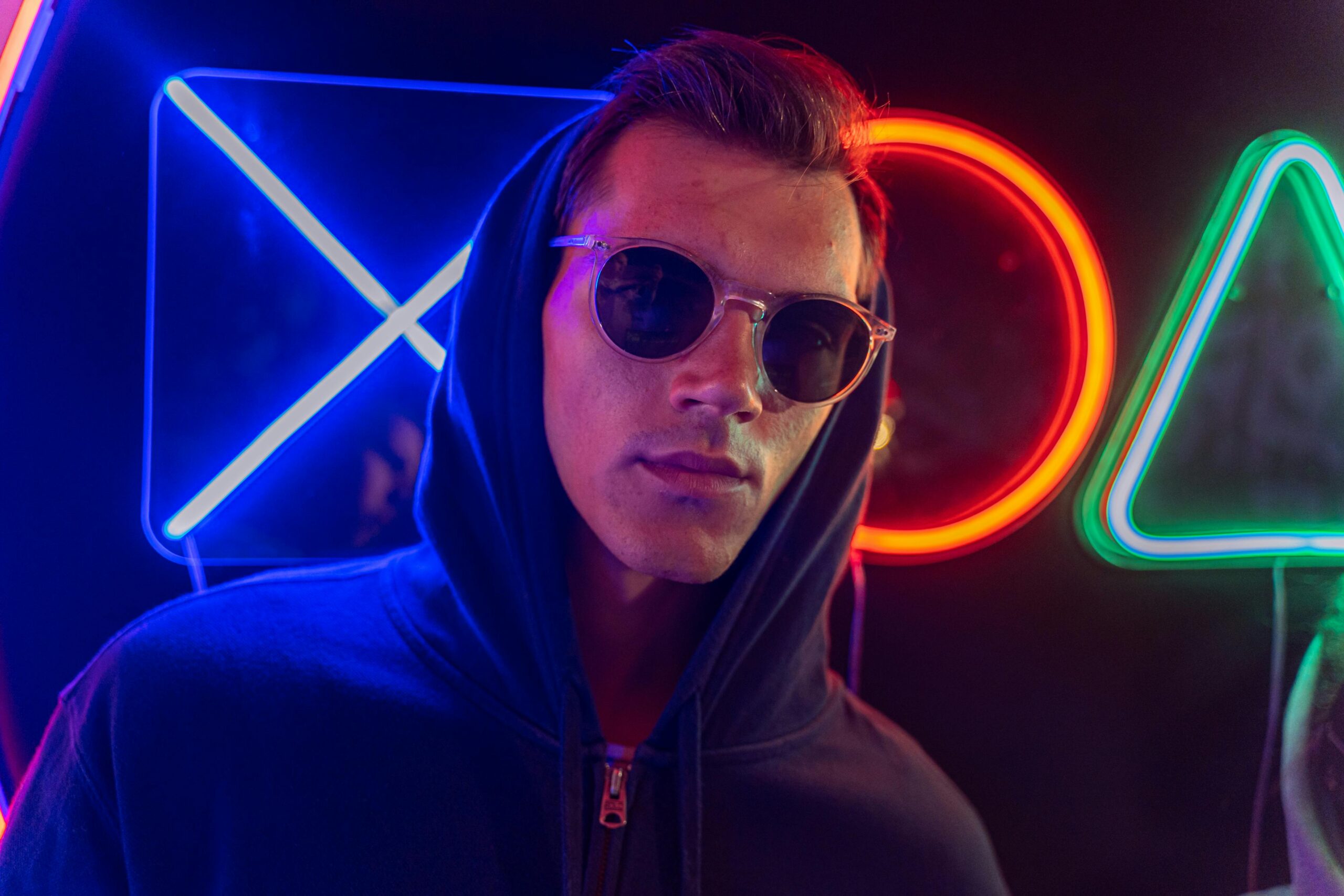 Man in sunglasses and hoodie with neon lights in the background.