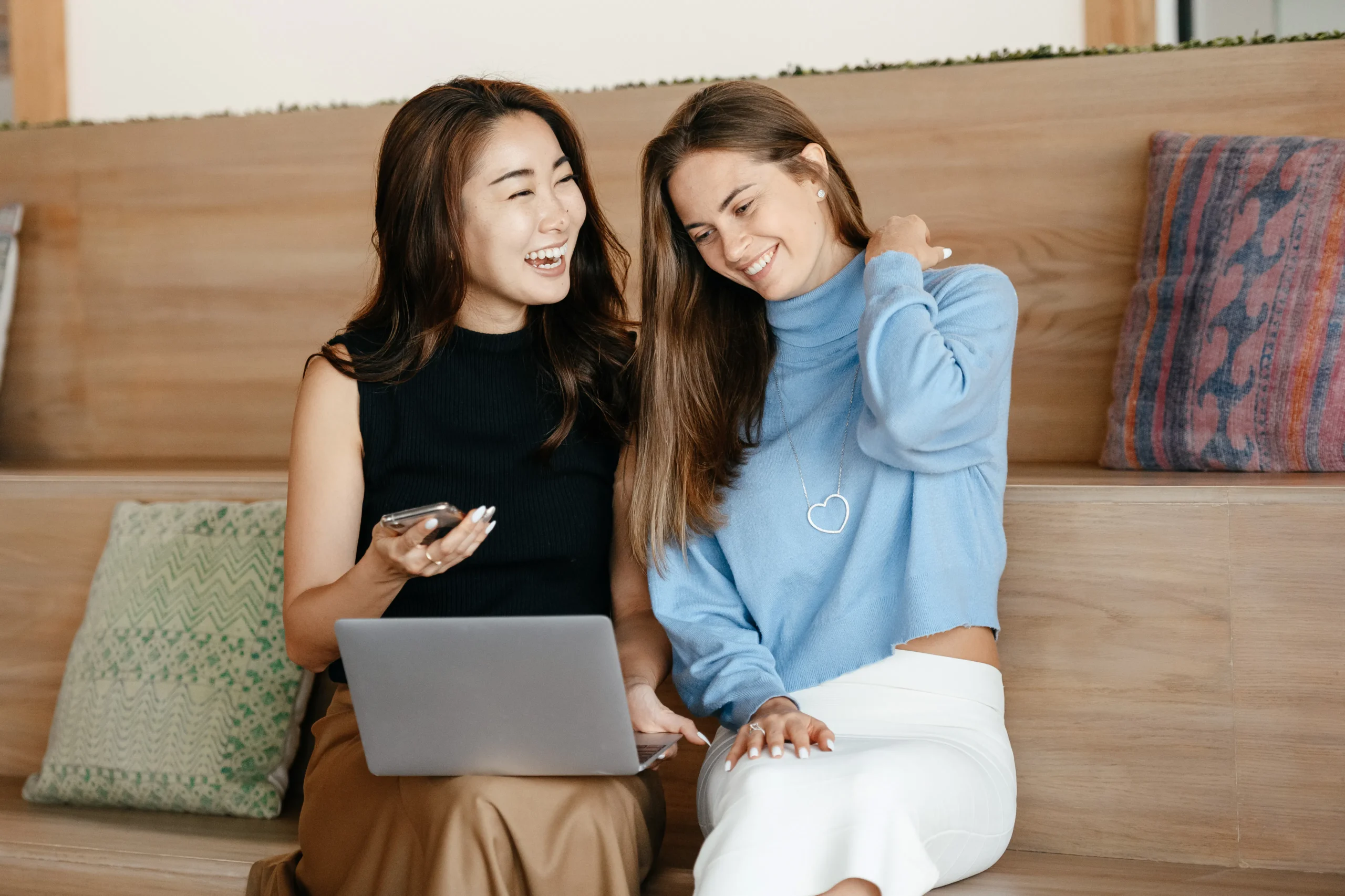 Two smiling women are collaborating on a laptop, possibly discussing native marketing strategies for effective native ads, while seated on a cushioned bench.